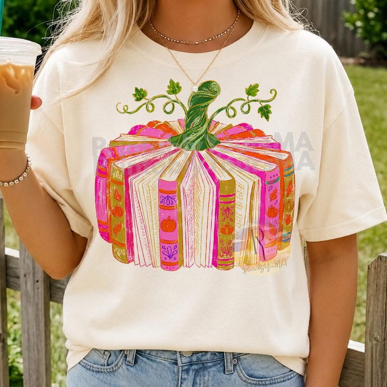May include: A cream-coloured t-shirt featuring a graphic of a pumpkin constructed from upright books. The books are shades of pink, orange, and gold with pumpkin illustrations on the spines. A green stem sits atop the book pumpkin design.