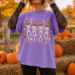 K&ouml;nnte beinhalten: Ein hellviolettes T-Shirt mit einer Grafik von vier Skeletten mit K&uuml;rbisk&ouml;pfen, die zwischen bunten Blumen und Bl&auml;ttern tanzen. Das T-Shirt hat kurze &Auml;rmel und wird mit schwarzen, transparenten Langarm-Elementen mit Spinnennetz-Muster getragen.
