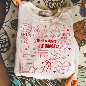 May include: A cream-colored sweatshirt with red graphic designs. The design includes hearts, bows, a key, and the words "Just Sold," "Dream Home," and "Home Sweet Home." The phrase "Home is Where the Heart Is" is also featured.