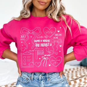 May include: A bright pink sweatshirt with white graphic text and illustrations related to real estate. The design includes hearts, bows, a key, and the words "Just Sold," "Sold," and "Home is Where the Heart Is."