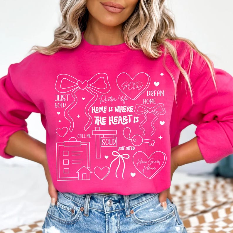 May include: A bright pink sweatshirt with white graphic text and illustrations related to real estate. The design includes hearts, bows, a key, and the words "Just Sold," "Sold," and "Home is Where the Heart Is."