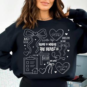 May include: Black sweatshirt with white graphic elements related to real estate. The design includes bows, hearts, a key, a house, and the words "Just Sold," "Dream Home," and "Home Sweet Home." The phrase "Home is Where The Heart Is" is also featured.