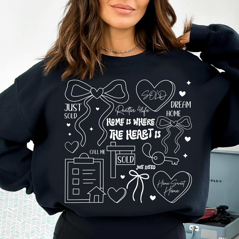 May include: Black sweatshirt with white graphic elements related to real estate. The design includes bows, hearts, a key, a house, and the words "Just Sold," "Dream Home," and "Home Sweet Home." The phrase "Home is Where The Heart Is" is also featured.