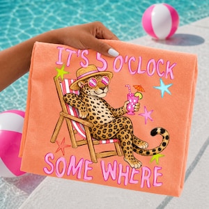 Op de afbeelding: Koraalroze strandlaken met een cartoon luipaard met zonnebril en strohoed, die een cocktail vasthoudt in een strandstoel. De tekst "IT'S 5 O'CLOCK SOME WHERE" staat in roze op de handdoek. Op de achtergrond zijn strandballen te zien.