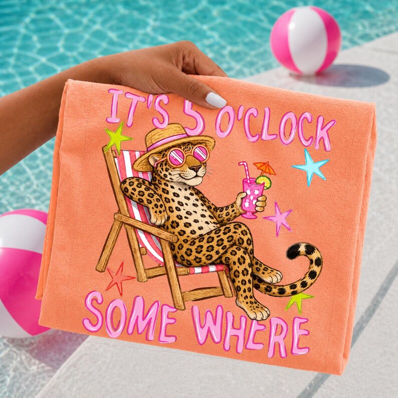 Op de afbeelding: Koraalroze strandlaken met een cartoon luipaard met zonnebril en strohoed, die een cocktail vasthoudt in een strandstoel. De tekst "IT'S 5 O'CLOCK SOME WHERE" staat in roze op de handdoek. Op de achtergrond zijn strandballen te zien.
