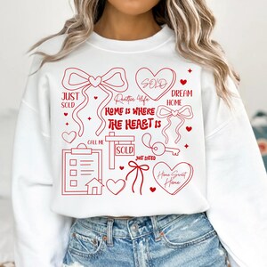 May include: White sweatshirt with red graphic elements related to real estate. The design includes bows, hearts, a key, and the phrases "Just Sold," "Home is Where the Heart Is," and "Home Sweet Home."