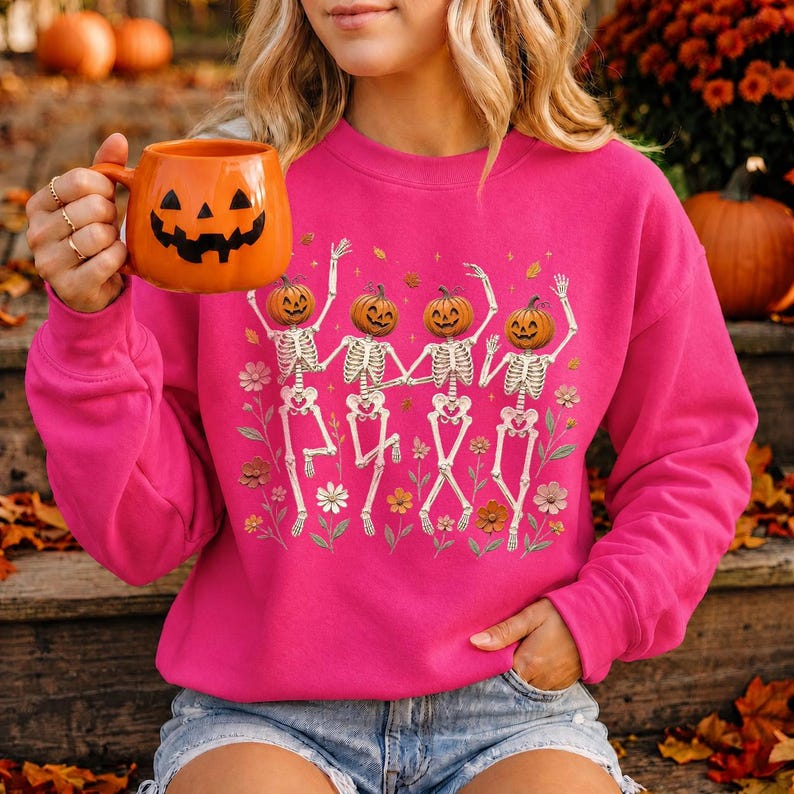 K&ouml;nnte beinhalten: Ein leuchtend pinkfarbenes Langarm-Sweatshirt mit einer Grafik von vier tanzenden Skeletten mit K&uuml;rbisk&ouml;pfen, umgeben von bunten Blumen. Das Sweatshirt wird mit einer leuchtend orangefarbenen Keramiktasse in K&uuml;rbisform kombiniert.