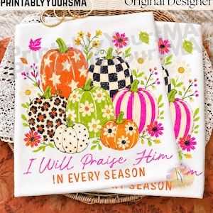 May include: A white t-shirt featuring a graphic of six pumpkins with various patterns including stars, leopard print, checkers, stripes, and flowers, surrounded by colorful floral accents. The text reads, I Will Praise Him IN EVERY SEASON.