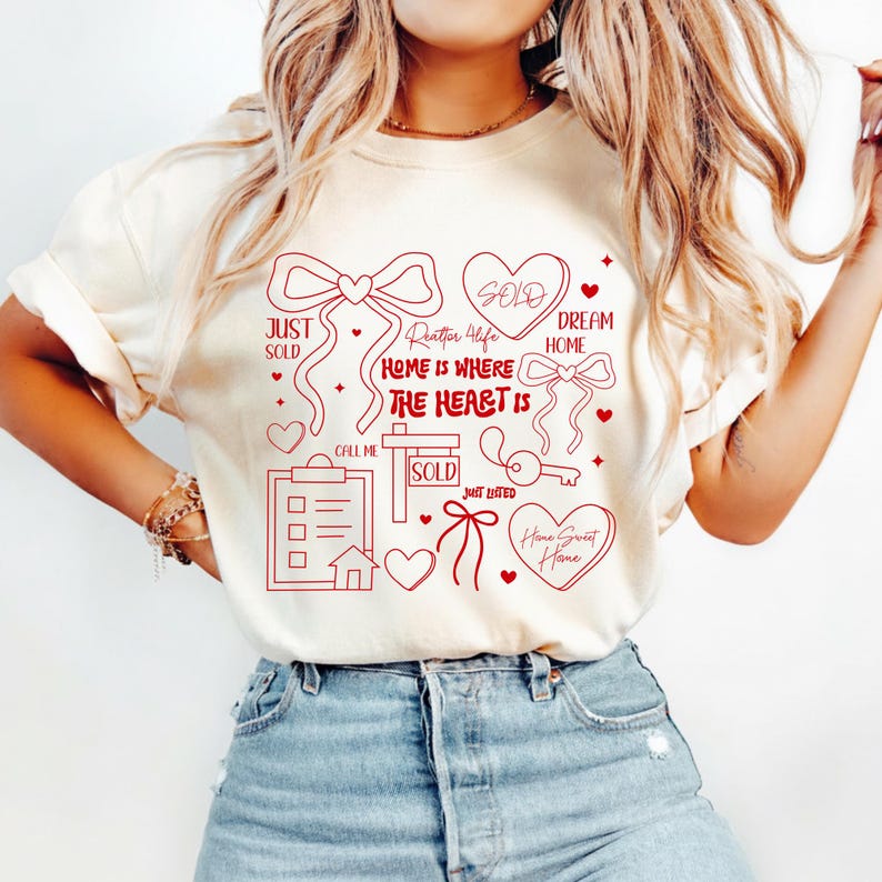 May include: Cream-colored t-shirt with red graphic elements related to real estate. The design includes bows, hearts, a key, a house, and the phrases "Just Sold," "Dream Home," and "Home Sweet Home."