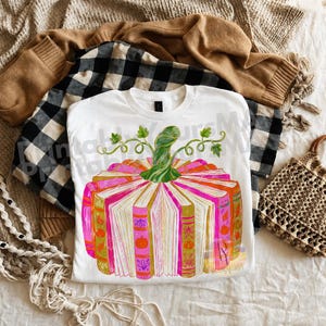 May include: A white t-shirt featuring a graphic of a pumpkin constructed from open books. The books have pink, orange, and gold spines with small pumpkin icons. A green stem with curling vines sits on top. The design includes a watermark text overlay.