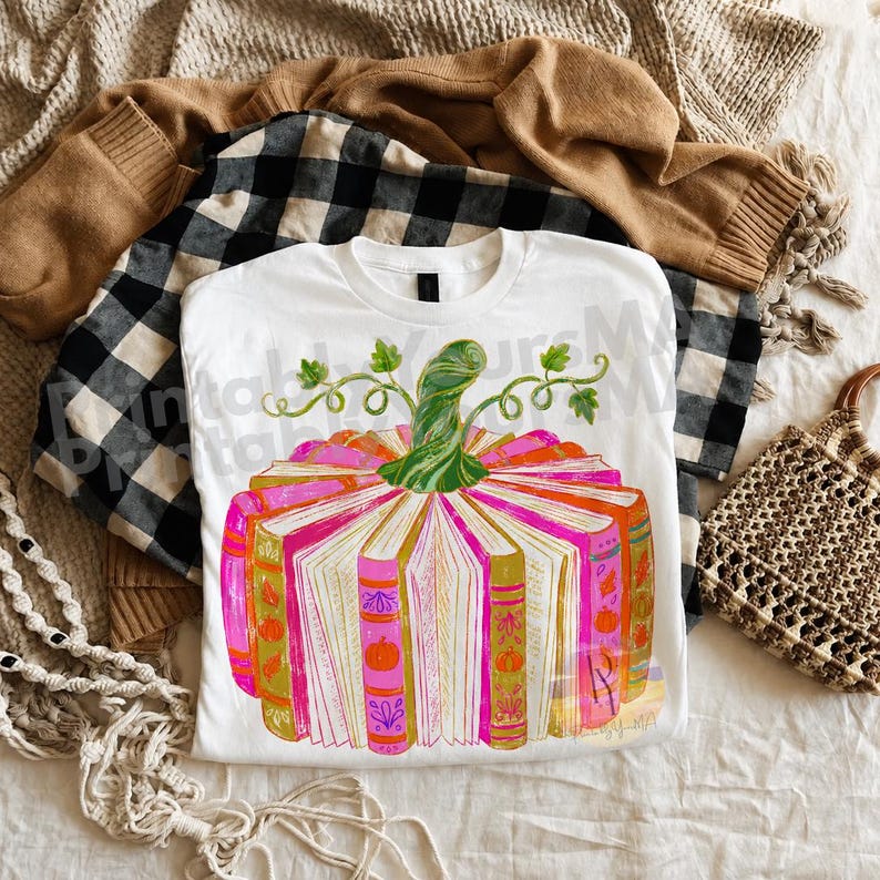 May include: A white t-shirt featuring a graphic of a pumpkin constructed from open books. The books have pink, orange, and gold spines with small pumpkin icons. A green stem with curling vines sits on top. The design includes a watermark text overlay.