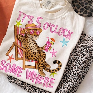 Op de afbeelding: Cr&egrave;mekleurig T-shirt met een luipaard met zonnebril en hoed, met een roze drankje. De tekst luidt "It's 5 O'Clock Somewhere" in roze. Een luipaardprint broek is zichtbaar.