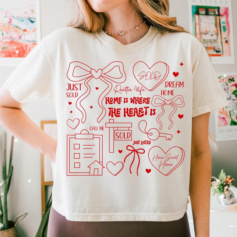 May include: Cream-colored t-shirt with a red graphic design. The design includes hearts, bows, a key, and the phrases "Just Sold," "Dream Home," and "Home Sweet Home." The text "Realtor 4 Life" and "Home is Where the Heart Is" are also featured.