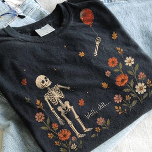 May include: A charcoal gray t-shirt featuring a graphic of a skeleton reaching for a red balloon. The design includes scattered autumn leaves, various orange, pink, and white flowers, and the printed text "Well, shit..." in a white script font.