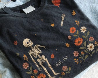 Może przedstawiać: Ciemnoszary t-shirt z grafiką przedstawiającą szkielet sięgający po czerwony balon. Wzór zawiera rozrzucone jesienne liście, różnorodne pomarańczowe, różowe i białe kwiaty oraz nadrukowany tekst "Well, shit..." białą, ozdobną czcionką.