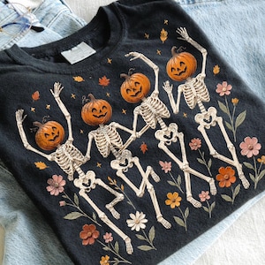 May include: A black t-shirt featuring a graphic print of three skeletons with jack-o'-lantern pumpkin heads. The skeletons are depicted in dancing poses, surrounded by small orange and pink flowers, green leaves, and scattered autumn leaves.