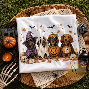 May include: A white t-shirt featuring a graphic of four dachshunds dressed in Halloween costumes. One wears a purple witch hat and cape, one is a ghost, one is inside a pumpkin, and one wears a vampire cape. Bats and autumn leaves surround them.