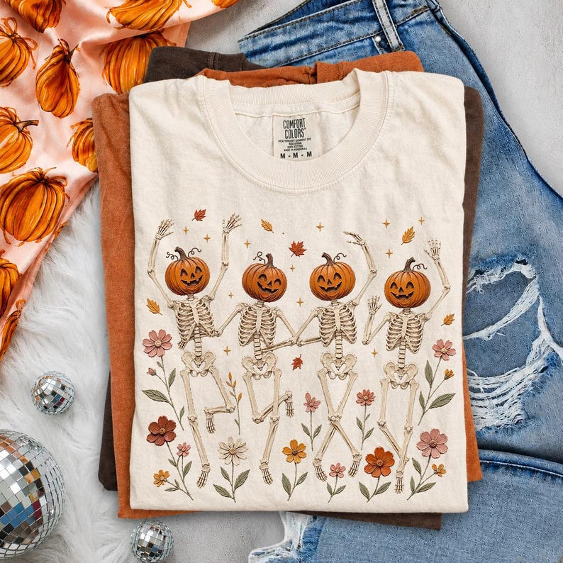 K&ouml;nnte beinhalten: Ein cremefarbenes Kurzarm-T-Shirt mit einer Grafik von vier Skeletten mit orangefarbenen K&uuml;rbisk&ouml;pfen, die zwischen bunten Blumen und fallenden Bl&auml;ttern tanzen. Das T-Shirt hat einen Rundhalsausschnitt und ein Comfort Colors-Etikett am Innenhals.