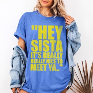 May include: A blue short-sleeved t-shirt featuring the yellow text "HEY SISTA IT'S REALLY REALLY NICE TO MEET YA.." printed on the front. The shirt is worn with a light blue denim jacket draped over the shoulders, showcasing a casual layered look.