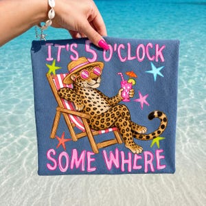 Op de afbeelding: Een blauwe strandlaken met een cartoon luipaard met een zonnebril en een strohoed, die een cocktail vasthoudt in een strandstoel. De tekst "IT'S 5 O'CLOCK SOME WHERE" staat in het roze op de handdoek.
