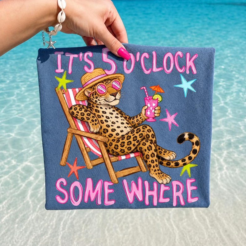 Op de afbeelding: Een blauwe strandlaken met een cartoon luipaard met een zonnebril en een strohoed, die een cocktail vasthoudt in een strandstoel. De tekst "IT'S 5 O'CLOCK SOME WHERE" staat in het roze op de handdoek.
