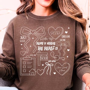 May include: Brown sweatshirt with white graphic elements related to real estate. The design includes hearts, bows, a key, a house, and the words "SOLD", "JUST SOLD", "DREAM HOME", "HOME IS WHERE THE HEART IS", "JUST LISTED", "Home Sweet Home", and "Realtor 4 Life".