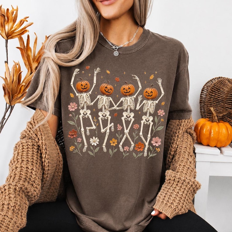 K&ouml;nnte beinhalten: Ein dunkelbraunes Kurzarm-T-Shirt mit einem Grafikdruck von vier Skeletten mit K&uuml;rbisk&ouml;pfen, die zwischen bunten Blumen und fallenden Bl&auml;ttern tanzen. Die Skelette sind in einer Reihe angeordnet, jedes mit erhobenen Armen und bewegten Beinen.