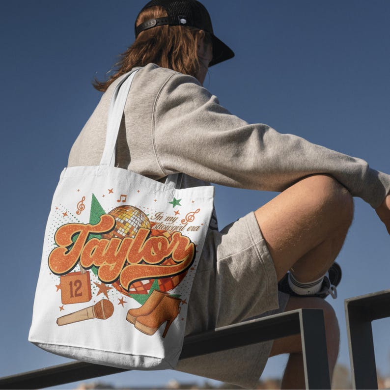 K&ouml;nnte beinhalten: Wei&szlig;e Tragetasche mit Retro-Design. Sie zeigt den Namen "Taylor" in Orange und Braun, eine Discokugel und Noten. Die Tasche enth&auml;lt auch den Text "In my 1989 girl era" und braune Stiefel.