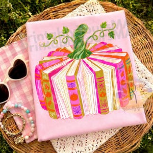 May include: A light pink t-shirt featuring a graphic of a pumpkin constructed from open books. The pumpkin has a green stem and leaves, with book spines in shades of orange, pink, and gold. The design includes small pumpkin and leaf icons.