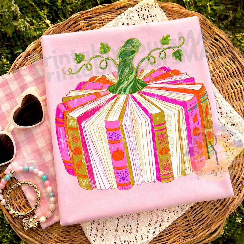 May include: A light pink t-shirt featuring a graphic of a pumpkin constructed from open books. The pumpkin has a green stem and leaves, with book spines in shades of orange, pink, and gold. The design includes small pumpkin and leaf icons.