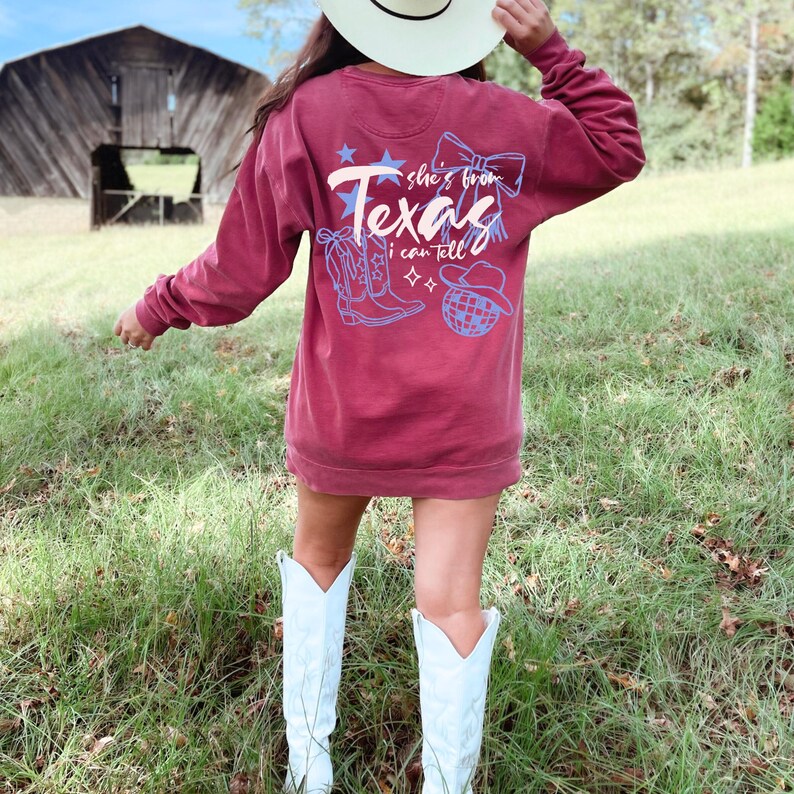 May include: Burgundy crewneck sweatshirt with the text "Texas" and illustrations of a cowboy boot, hat, and disco ball. The sweatshirt has long sleeves and is worn with white cowboy boots. The background includes a barn and green grass.