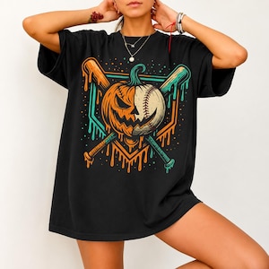 Może przedstawiać: Czarny t-shirt z kr&oacute;tkim rękawem z nadrukiem graficznym przedstawiającym dynię przeciętą na p&oacute;ł, z jedną stroną ukazującą wyrzeźbioną twarz dyni, a drugą przypominającą piłkę baseballową, skrzyżowaną z dwoma kijami baseballowymi w kolorze pomarańczowym i morskim.
