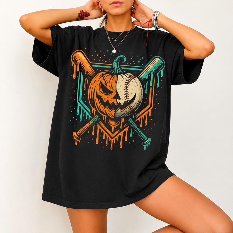 Może przedstawiać: Czarny t-shirt z kr&oacute;tkim rękawem z nadrukiem graficznym przedstawiającym dynię przeciętą na p&oacute;ł, z jedną stroną ukazującą wyrzeźbioną twarz dyni, a drugą przypominającą piłkę baseballową, skrzyżowaną z dwoma kijami baseballowymi w kolorze pomarańczowym i morskim.
