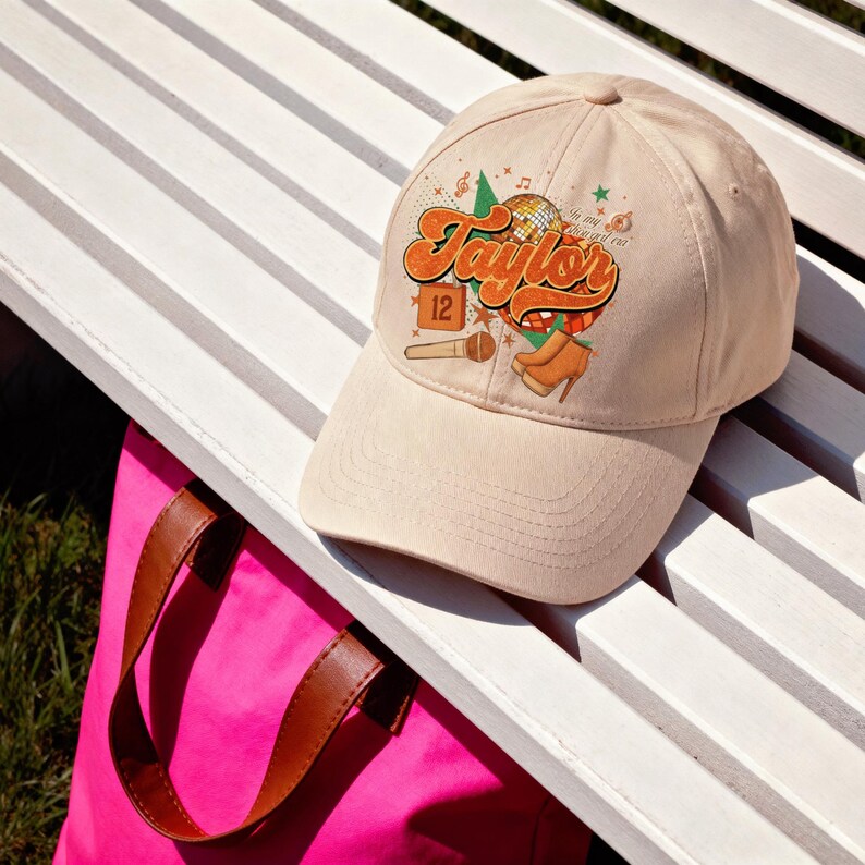 K&ouml;nnte beinhalten: Beige Baseballkappe mit dem Namen "Taylor" in orangefarbener Schrift, einer Discokugel und einer Mikrofon-Grafik. Die Zahl "12" ist ebenfalls abgebildet. Die Kappe liegt auf einer wei&szlig;en Bank neben einer pinkfarbenen Tragetasche.