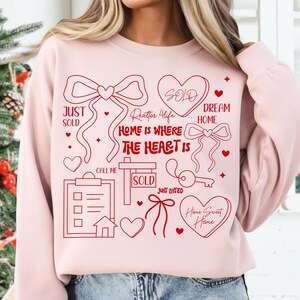 May include: A light pink sweatshirt with a red graphic design. The design includes hearts, bows, and the phrases "Home is Where the Heart Is", "Just Sold", "Dream Home", and "Home Sweet Home".