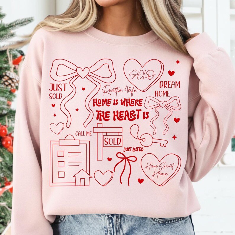 May include: A light pink sweatshirt with a red graphic design. The design includes hearts, bows, and the phrases "Home is Where the Heart Is", "Just Sold", "Dream Home", and "Home Sweet Home".