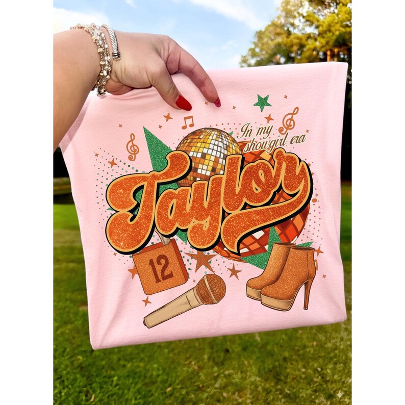 K&ouml;nnte beinhalten: Hellrosa T-Shirt mit Retro-Design. Das Design zeigt den Namen "Taylor" in Orange, eine Discokugel und braune Stiefel. Ein Mikrofon, die Zahl 12 und der Satz "In my showgirl era" sind ebenfalls abgebildet.