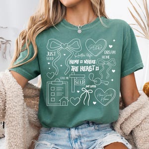 May include: Green t-shirt with white graphic elements related to real estate. The design includes hearts, bows, a key, and the words "SOLD", "Just Sold", "Dream Home", and "Home Sweet Home". The phrase "Home is where the heart is" is also featured.
