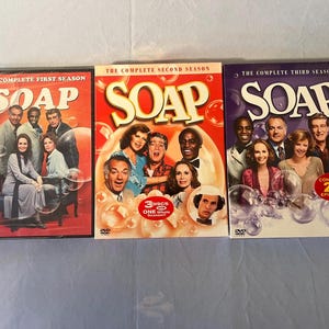 Puede incluir: Tres cajas de DVD de la serie de televisión "SOAP". La primera temporada está en una caja roja, la segunda en una caja roja y la tercera en una caja morada. Cada caja presenta una foto del elenco y el título del programa.