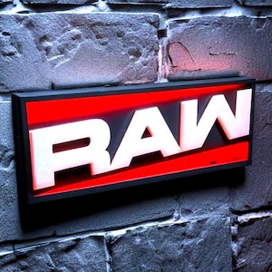 May include: A rectangular sign with the word "RAW" in bold, white letters against a red background. The sign is mounted on a gray brick wall, illuminated with a soft glow.