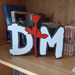 Op de afbeelding: Een wit en zwart 3D-geprint beeldhouwwerk van de letters "DM" met een rode roos in het midden, tentoongesteld op een houten plank. Het beeldhouwwerk is omgeven door boeken en manga.