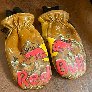Puede incluir: Un par de manoplas de cuero color canela Kinco con una ilustración gráfica de un toro rojo, un sol amarillo, picos de montañas y copos de nieve. El texto Red Bull está impreso en letras rojas grandes en la parte inferior de cada manopla.