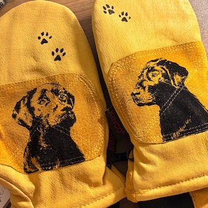 Puede incluir: Un par de manoplas de cuero amarillo con un estampado gráfico negro de una cabeza de perro en cada manopla. Sobre el gráfico del perro hay dos pequeñas huellas negras. Una etiqueta negra con el texto KINCO es visible en el lateral derecho.