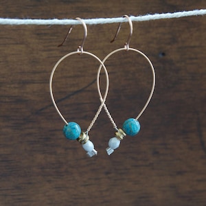May include: A pair of gold-colored hoop earrings with turquoise beads and small white and silver accents. The earrings are suspended against a dark wood background, showcasing their delicate design and color contrast.