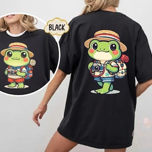 Może przedstawiać: Czarny t-shirt z nadrukiem kreskówkowej żaby. Żaba ma na sobie słomkowy kapelusz, szorty i plecak, trzyma aparat fotograficzny. Żaba jest zielona z różowymi policzkami. Projekt znajduje się również na plecach koszulki.