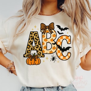 Peut inclure: T-shirt crème avec les lettres "ABC" sur le thème d'Halloween. La lettre "A" a un motif léopard, "B" est rempli de citrouilles et "C" a un motif à carreaux orange et blanc. Comprend des chauves-souris, une araignée et une citrouille.