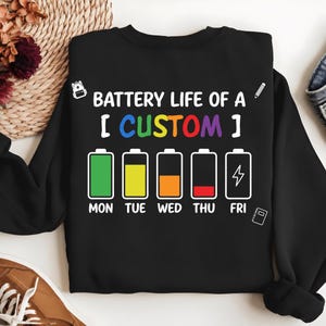 May include: A black long-sleeve sweatshirt featuring a graphic with the text BATTERY LIFE OF A [ CUSTOM ] and five battery icons representing Monday through Friday with varying charge levels in green, yellow, orange, red, and white.