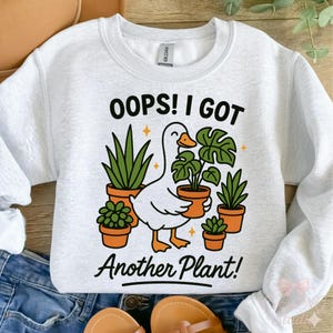 May include: A light gray sweatshirt with the text "OOPS! I GOT Another Plant!" and a cartoon goose holding a potted plant. The goose is surrounded by other potted plants in orange pots. The design is in black, orange, green, and white.