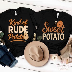 May include: Two black t-shirts with orange text and potato graphics. One shirt says "Kind of RUDE Potato" with an angry potato. The other says "Sweet POTATO" with a smiling potato. A tan hat sits below the shirts.