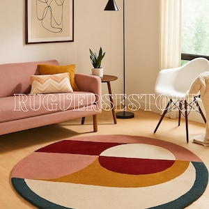 Puede incluir: Una alfombra redonda con un diseño geométrico. La alfombra presenta un patrón de bloques de color con secciones rosas, rojas, amarillas, crema y turquesas. Está sobre un suelo de madera en un salón.
